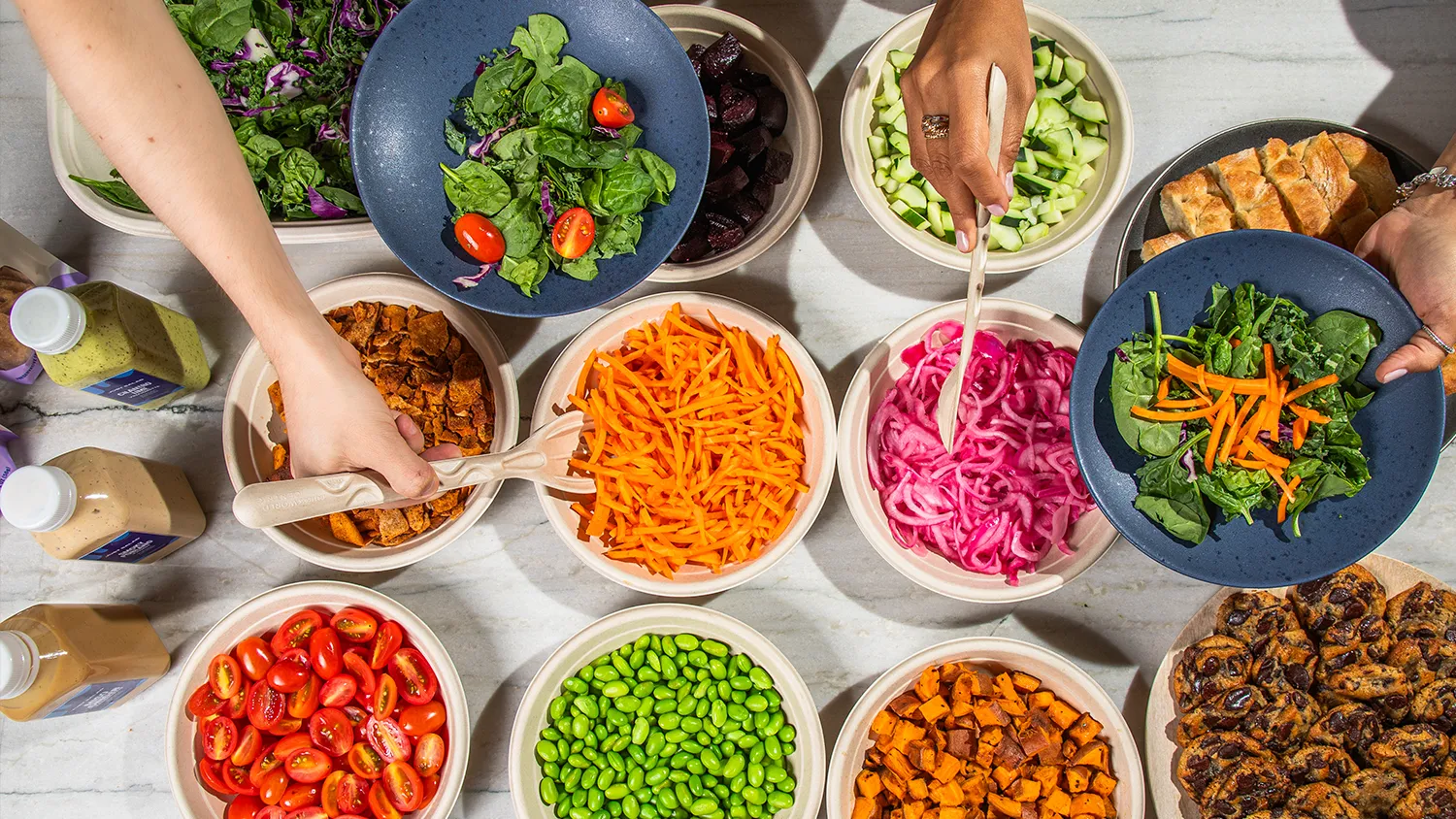 Overhead view of bowls with fresh salad ingredients including spinach, cherry tomatoes, shredded carrots, cubed cucumbers, beets, edamame, sweet potatoes, pickled onions, and bread with two hands preparing salads.