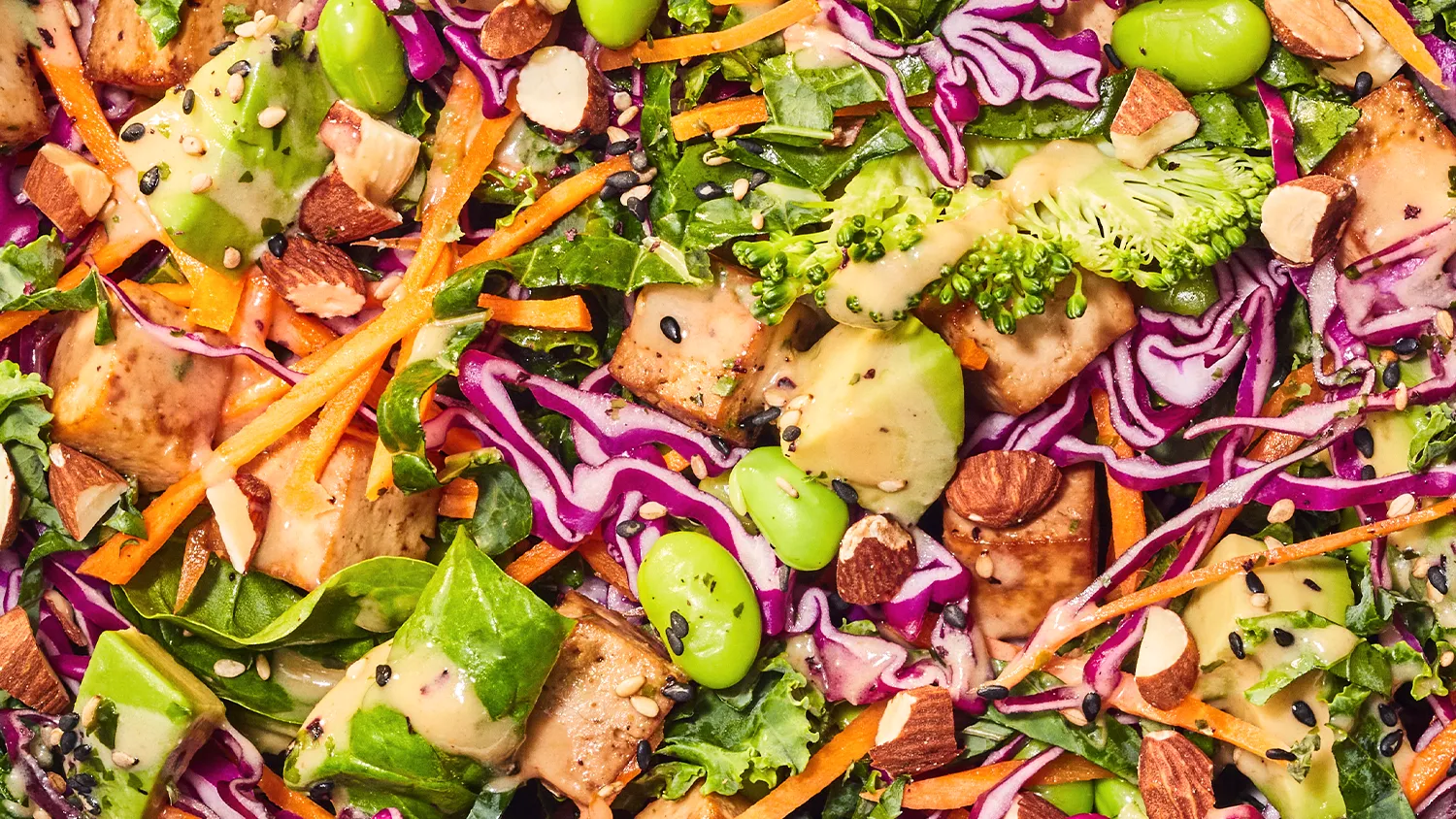 Colorful salad with broccoli, purple cabbage, carrot strips, tofu cubes, almonds, edamame, spinach, and creamy dressing with sesame seeds.