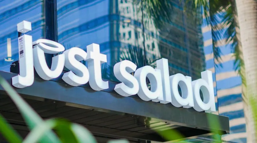 White 3D sign reading 'just salad' mounted on a building with glass windows reflecting nearby structures and palm trees.