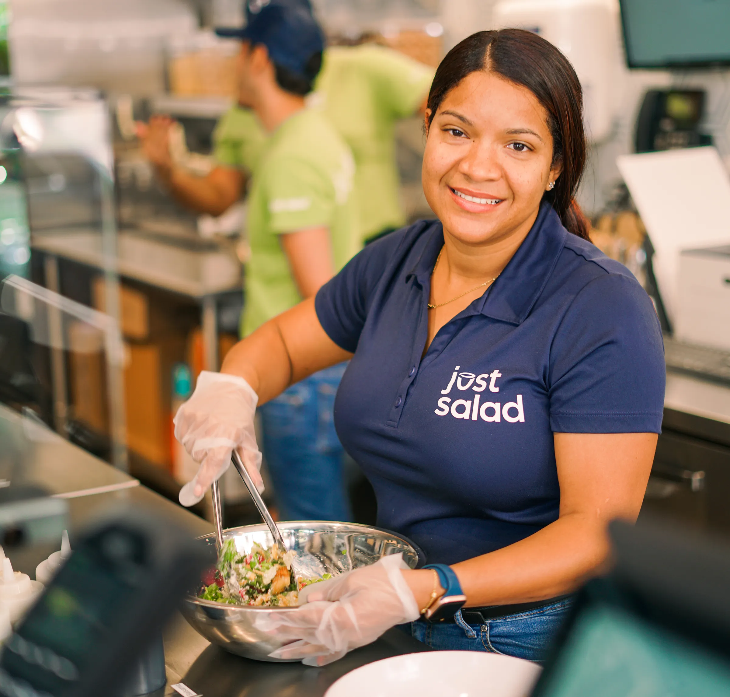 Smiling woman wearing a Just Salad uniform preparing a salad in a metal bowl with tongs inside a restaurant kitchen.