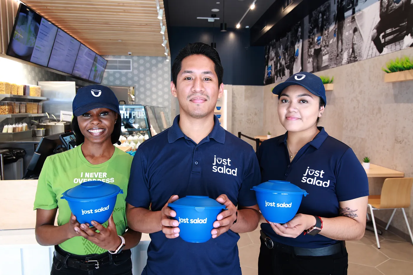 Three Just Salad employees in uniforms holding blue Just Salad bowls inside a restaurant.