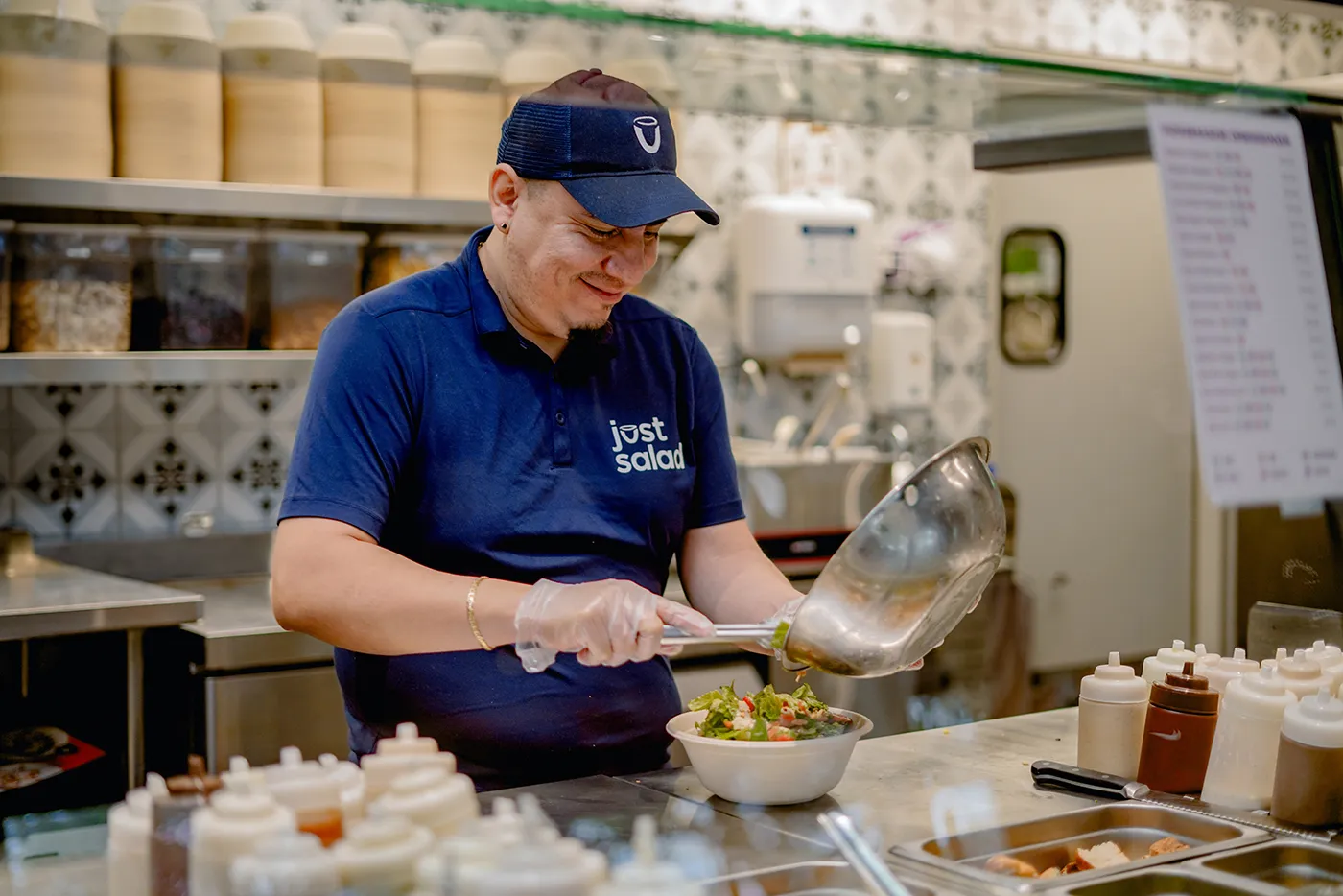 Smiling man wearing blue Just Salad uniform preparing a salad bowl in a kitchen.