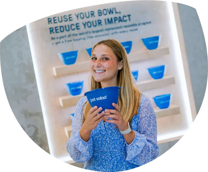 Smiling woman holding a blue reusable bowl in front of a display with several similar blue bowls and the text 'Reuse your bowl, reduce your impact.'