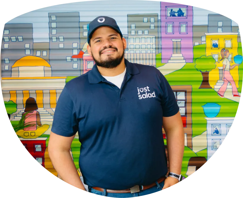 Smiling man wearing a navy Just Salad polo shirt and cap standing in front of a colorful cityscape mural.