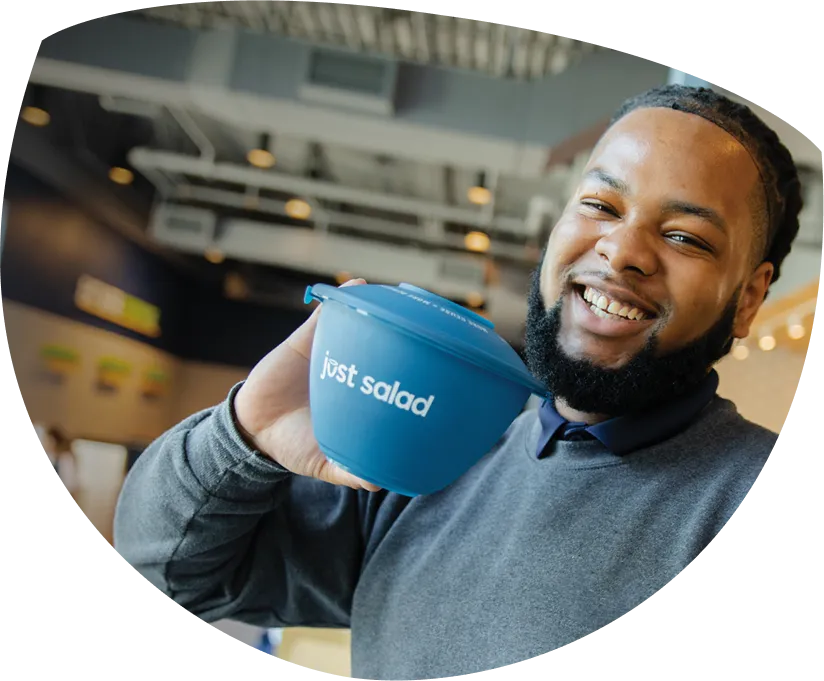 Smiling man holding a blue bowl with the 'just salad' logo inside a modern restaurant.